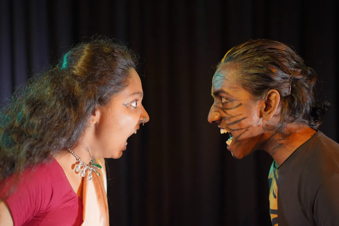 Two actors face each other mid-scene, faces painted, in a Nirmik production.